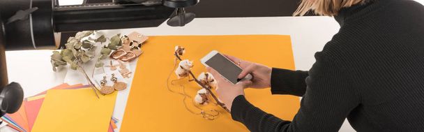 Ausgeschnittene Ansicht des Fotografen, der Komposition mit Baumwollblume und Accessoires auf Smartphone fotografiert, Panoramaaufnahme - Foto, Bild