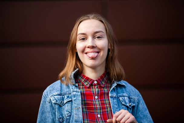 Emotions. Happy. Teenager. Young pretty white caucasian girl in casual jean jacket shows tongue opposite dark modern backgroun on the street in the autumn. - Foto, imagen