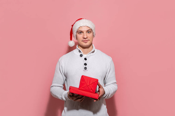 Handsome man in a white sweater and red Santa hat holding Christmas gifts in red boxes isolated on pink background. - Фото, зображення