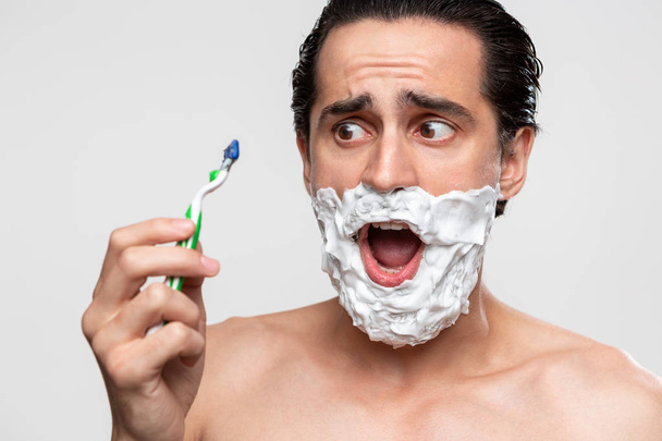 Красивий чоловік з вусами використовує палицю для гоління та піну для гоління, щоб обрізати бороду, що стоїть оголеною, ізольовано на білому тлі
. - Фото, зображення
