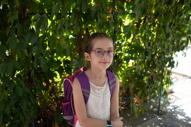 Schulmädchen mit violettem Rucksack und lila Brille. Mädchen im weißen Kleid, trägt im Herbst eine Brille. - Foto, Bild