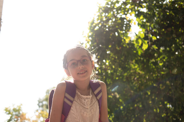 Iskoláslány lila hátizsákkal és lila szemüveggel. Fehér ruhás lány, ősszel szemüvegben.. - Fotó, kép