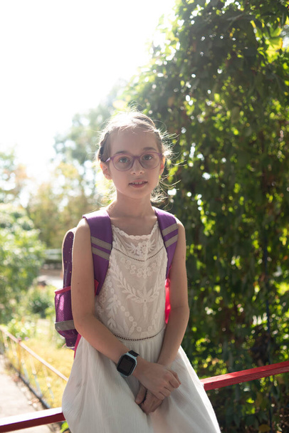 Koulutyttö violetti reppu ja violetti lasit. Tyttö valkoisessa mekossa, silmälasit syksyllä
. - Valokuva, kuva