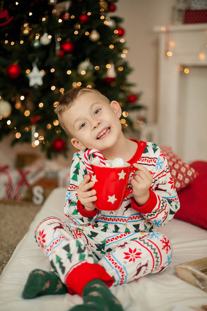 Ένα αγοράκι με χριστουγεννιάτικες πιτζάμες κάθεται κοντά σε ένα χριστουγεννιάτικο δέντρο και κρατάει μια κούπα κακάο με ζαχαρωτά. Χριστούγεννα. Νέο έτος. - Φωτογραφία, εικόνα