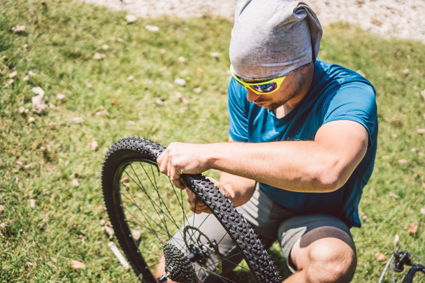 自転車修理。マウンテンバイクを修理する男。事故のトラブル後輪ケースにサイクリストの男。男はイタリアの背景の山の湖の近くの自転車を修正します。自転車を屋外で修理するサイクリスト - 写真・画像