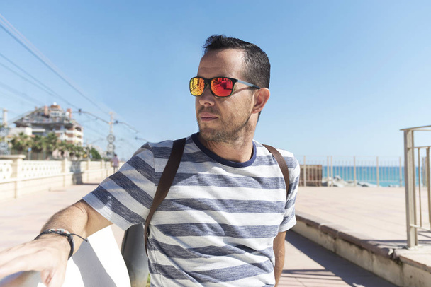 Młody brodaty mężczyzna w okularach przeciwsłonecznych siedzący na promenadzie odwracając wzrok - Zdjęcie, obraz