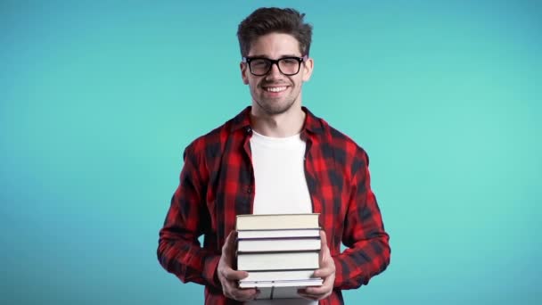 European student in red plaid shirt on blue background in studio holds stack of university books from library. Guy smiles, he is happy to graduate. - Video