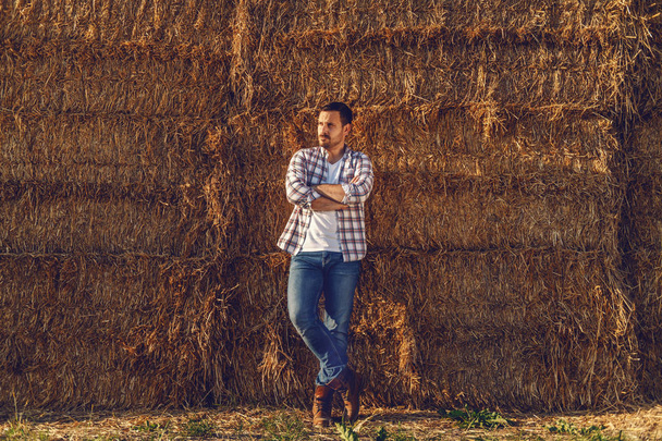 Attractive caucasian young farmer leaning on bales of hay and holding arms crossed while looking away. - Foto, Bild