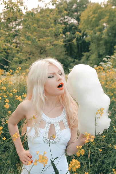 Charming adult blonde girl eating cotton candy on the nature in the park alone - Photo, Image