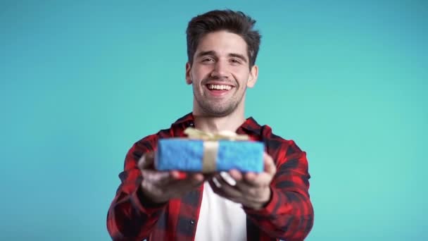 Handsome man holding christmas gift on blue studio background smiles to camera. Happy european guy in red shirt. Slow motion. - Metraje, vídeo