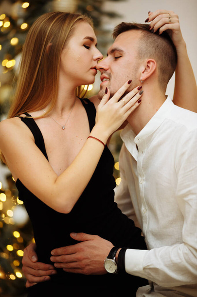 ζευγάρι στην αγάπη φιλιά και αγκαλιές στον καναπέ κοντά στα φώτα χριστουγεννιάτικο δέντρο. Νύχτα Πρωτοχρονιάς. Χριστούγεννα. - Φωτογραφία, εικόνα