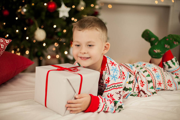 Маленький хлопчик у різдвяній піжамі сидить біля ялинки і відкриває подарунки. Різдво. Новий рік
. - Фото, зображення