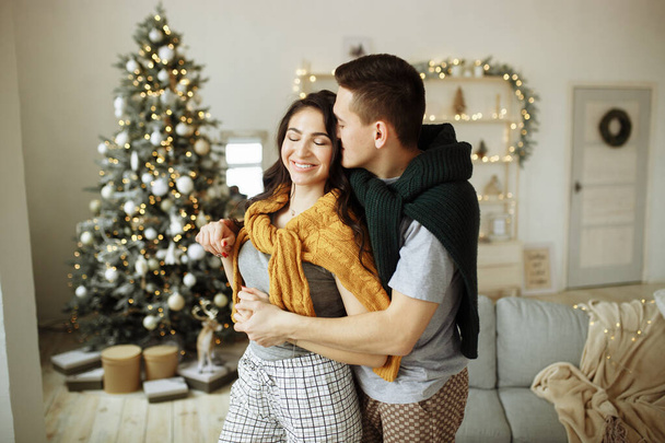 happy couple in love in the morning near the Christmas tree, a guy hugs a girl, morning Christmas. hearth, home clothes and sweaters. - Φωτογραφία, εικόνα