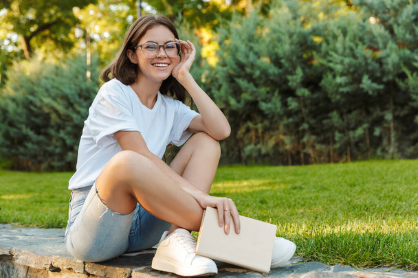 Smiling young girl holding a book while sitting in the park - Photo, Image
