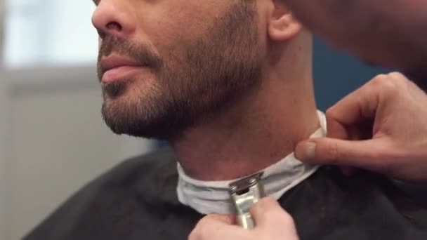Friseur rasiert den Kunden den Bart auf einem Stuhl. Bartschnitt. Friseur, um einen Bart mit einem elektrischen Rasierer zu rasieren. Pflege des echten Menschen. Seitenansicht eines jungen bärtigen Mannes, der beim Friseur einen Bartschnitt bekommt - Filmmaterial, Video