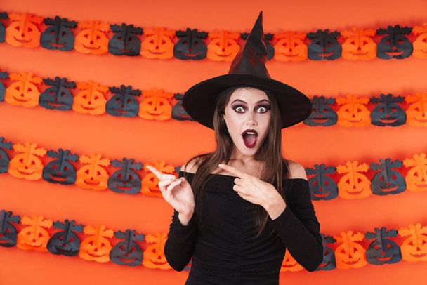 Image of shocked witch girl in black halloween costume pointing  - Foto, Imagem