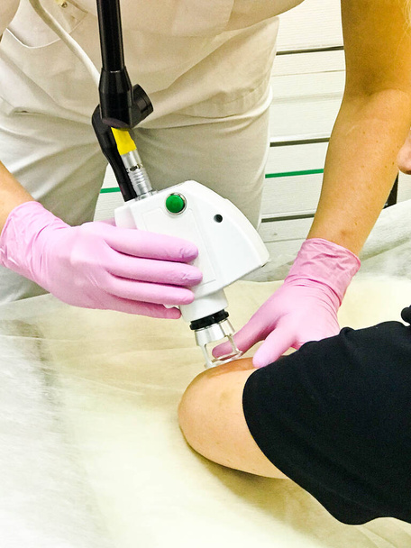 Un cosmétologue, médecin spécialiste, effectue une procédure de cosmétologie de rajeunissement laser des coudes au patient avec ses mains dans des gants de caoutchouc roses à l'aide de l'appareil dans le salon de beauté. - Photo, image