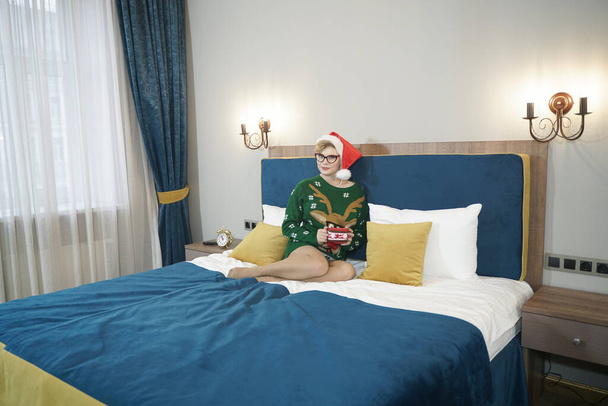 可愛いですプラスサイズオタク女の子で眼鏡と緑のクリスマスセーターとともにトナカイと赤サンタクロースの帽子で午前中にベッドの上にポーズ - 写真・画像