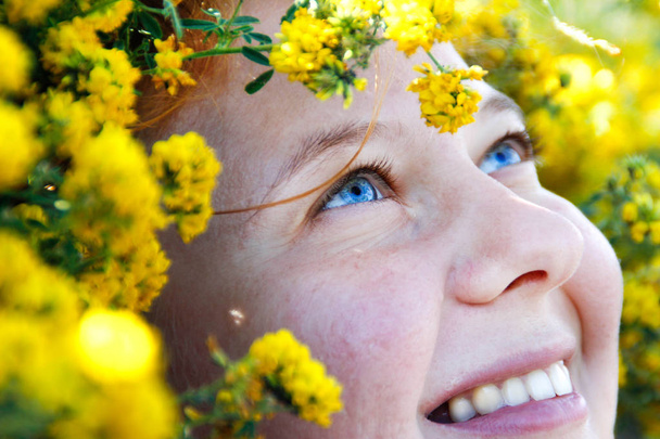 Um retrato muito próximo com ênfase nos incríveis olhos azuis de uma menina de cabelos vermelhos cercada por flores amarelas brilhantes. Sardas, rugas ao redor dos olhos e um sorriso alegre
. - Foto, Imagem