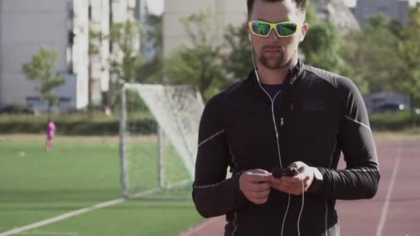 Tema spor ve sağlık. Kafkasyalı genç bir adam kulaklıkla müzik dinlemek için akıllı telefon akıllı fton kullanıyor. Şehir stadyumundaki koşu bandında teknoloji ve mobil uygulama çalma listesi - Video, Çekim