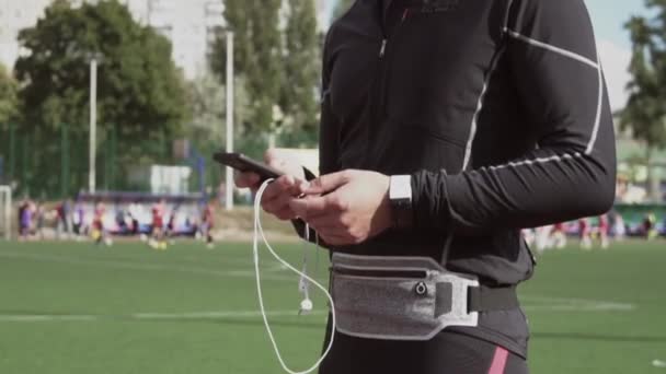 Theme sport and health. A young Caucasian man uses a smartphone smarfton to listen to music on headphones. Technology and mobile application playlist on the city stadium track treadmill - Footage, Video