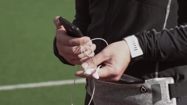 Tema deporte y salud. Un joven caucásico usa un smartphone smarfton para escuchar música en los auriculares. Lista de reproducción de tecnología y aplicaciones móviles en la cinta de correr del estadio de la ciudad
 - Metraje, vídeo