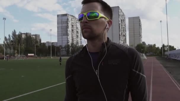 Themasport en gezondheid. Portret van een jonge blanke man die naar muziek luistert met een koptelefoon. Trainingssportles bij Stadstadionloopband - Video