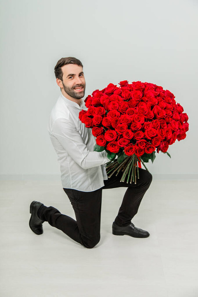 Full Length záběr pohledný mladý muž drží růže a dívá se na kameru, usmívá se při pokleknutí, na bílém pozadí. - Fotografie, Obrázek