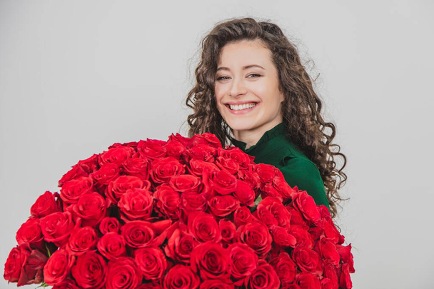Closeup portrait of a satisfied young woman dressed in green dress holding huge bouquet of roses isolated over white background. - Фото, зображення