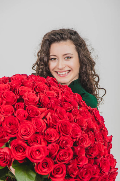 Closeup portrait of a satisfied young woman dressed in green dress holding huge bouquet of roses isolated over white background. - Фото, изображение