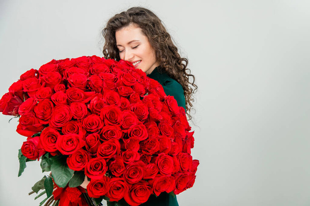 Cute curly brunette girl standing, smelling red roses over white background. - Fotó, kép