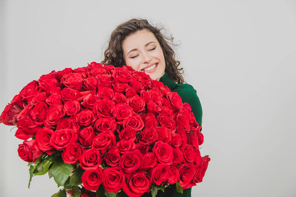 Cute curly brunette girl standing, smelling red roses over white background. - 写真・画像