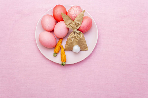 Paaseieren concept, tafelschikking decoratie. Roze (roze) eieren met konijn (konijn) papier cadeau ei wikkelen op plaat met gebreide wortelen. Laken achtergrond, bovenaanzicht. - Foto, afbeelding