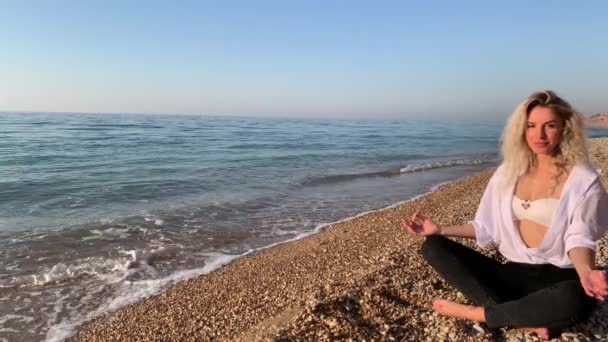 Hermosa mujer rubia sentada y meditando en la playa cerca de las olas del mar azul.Crimea, Sebastopol
. - Metraje, vídeo