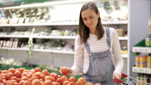 Young woman in denim overall is selecting fresh tomatoes at grocery store. - Кадры, видео