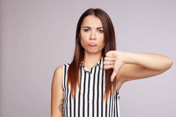 Irritated young exasperate  tan skinned woman keeps thumbs down, strongly dissatisfied with situation, has frowned eyebrows, demonstrates anger, wears casual blouse, stands against grey wall background - 写真・画像