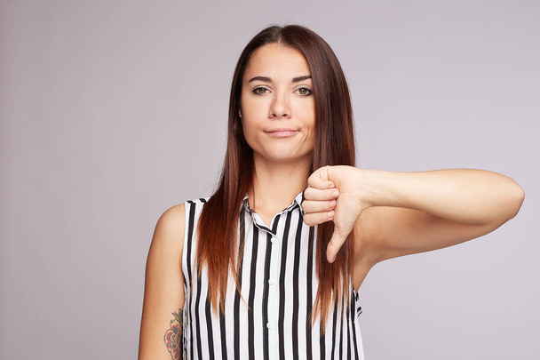 Discontent European woman shows disapproval sign, keeps thumb down, expresses dislike, frowns face in discontent, dressed in black and white shirt, isolated over pink background. Body language concept - Фото, зображення