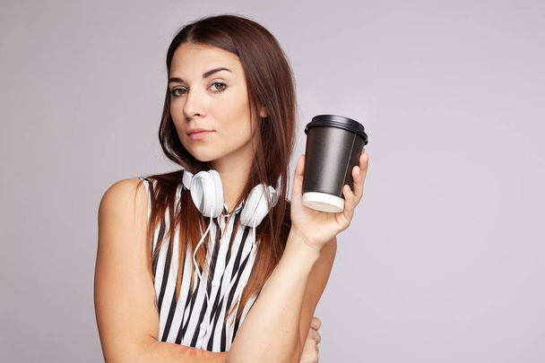 Time for music. Confident tan skinned female holds coffee to go in disposable cup, going to listen pleasant melody in headphones, looks flirty at camera, wears casual striped blouse, poses in studio. - Photo, image