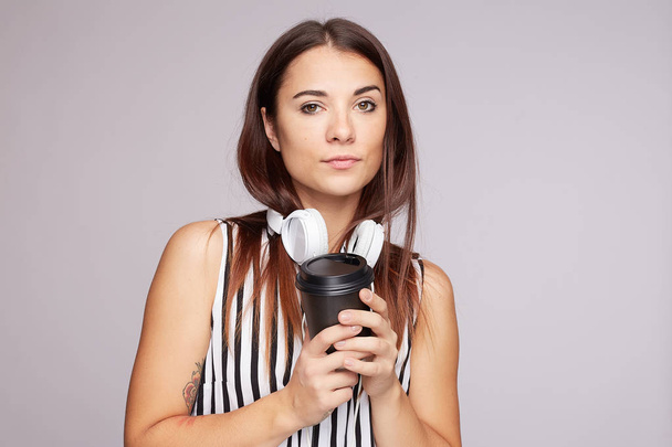 Komoly női diák tartja eldobható csésze mindkét kezében, fáradt kifejezés, megpróbál frissíteni erős forró kávé és zene, visel fekete-fehér blúz, áll beltéri szabad hely félre. - Fotó, kép