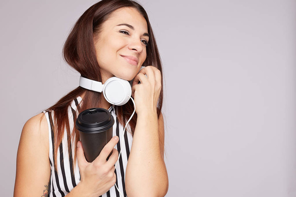 Izolovaný studiový záběr šťastné dámy v pohybu drží jednorázový šálek, pije kávu, poslouchá příjemnou hudbu ve sluchátkách, má volný čas, nosí pruhovanou blůzu, jemně se usmívá, má nejoblíbenější aktivitu. - Fotografie, Obrázek