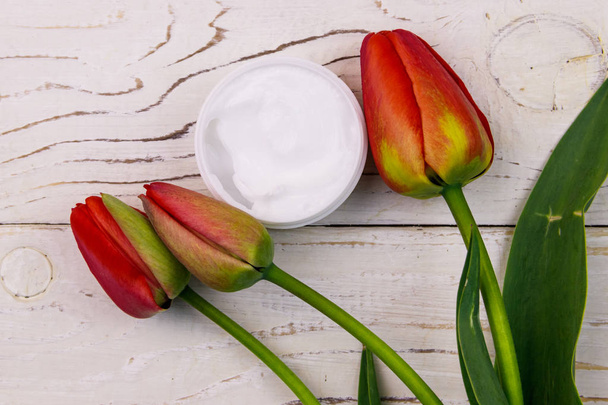 Sklenice hydratační krém s červenými tulipány na bílém dřevěném pozadí. Horní pohled - Fotografie, Obrázek