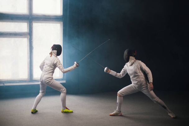 A fencing training in the studio - two women in white protective costumes having a duel - Foto, afbeelding