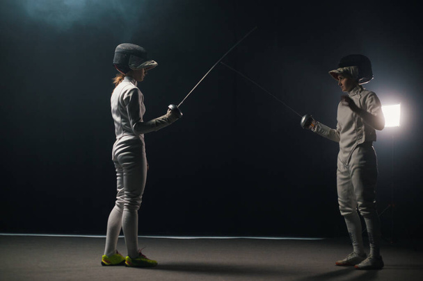 Две молодые женщины, тренирующиеся в фехтовальном поединке в дымчатой студии - приветствуют друг друга и начинают поединок
 - Фото, изображение