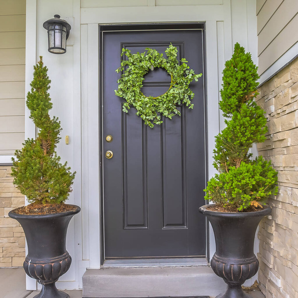 Квадратная Серая входная дверь дома с зеленым венком и окруженная высокими горшечными растениями
 - Фото, изображение