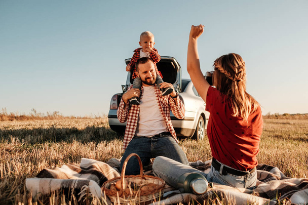 Happy Young Family Mom and Dad with Their Little Son Enjoying Summer Weekend Picnic Sitting on the Plaid Near the Car Outside the City in the Field at Sunny Day Sunset, Vacation and Road Trip Concept - Photo, Image