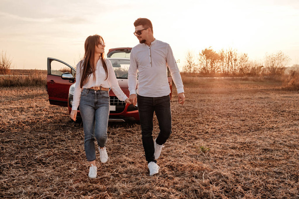 Beyaz gömlekli ve kot pantolonlu genç mutlu çift yeni arabalarında yolculuğun tadını çıkarıyor, Sahada güzel bir gün batımı, tatil ve seyahat konsepti - Fotoğraf, Görsel