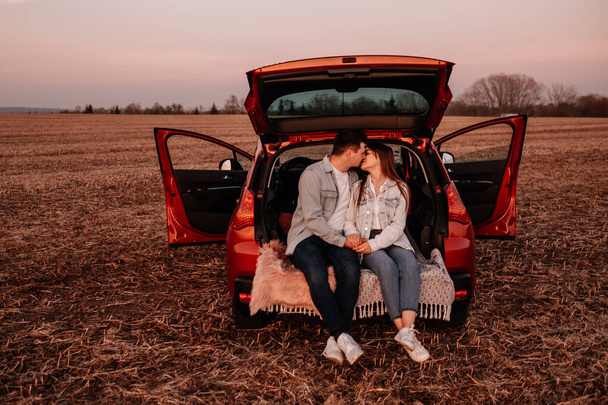 Fiatal boldog pár öltözött Alike fehér ing és farmer ül az új autó csomagtartó, gyönyörű naplemente a pályán, nyaralás és utazási koncepció - Fotó, kép