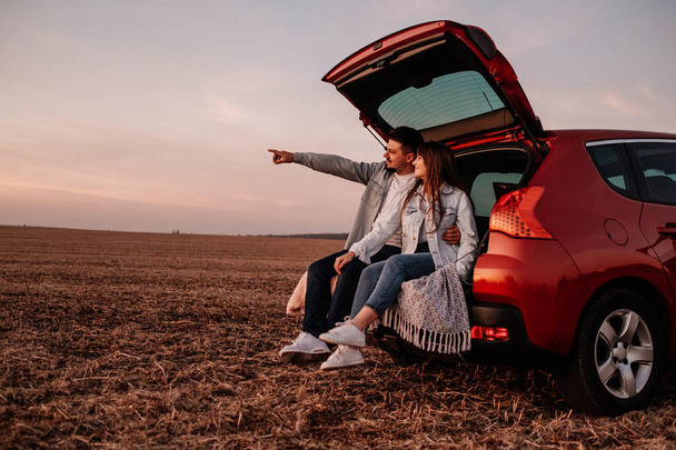 Joven pareja feliz vestida como en camisa blanca y pantalones vaqueros sentados en su nuevo baúl de coche, hermoso atardecer en el campo, vacaciones y concepto de viaje
 - Foto, imagen