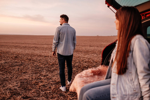 Young Happy Couple Dressed Alike in White Shirt and Jeans Sitting at Their New Car Trunk, Beautiful Sunset on the Field, Vacation and Travel Concept - Фото, зображення