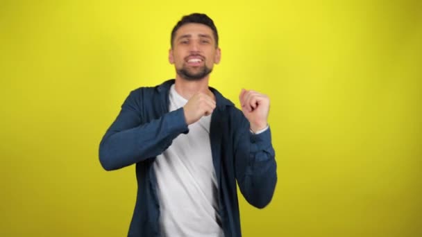 A young man in a white T-shirt and blue shirt is dancing with a smile on a yellow background with copy space. 4k footage - Footage, Video